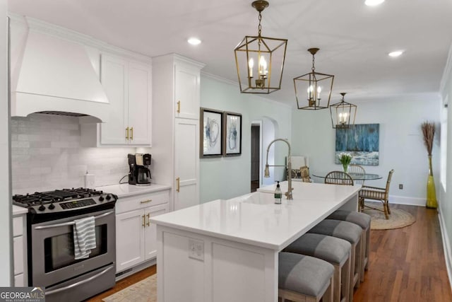 kitchen featuring pendant lighting, custom exhaust hood, stainless steel gas range oven, a center island with sink, and white cabinets