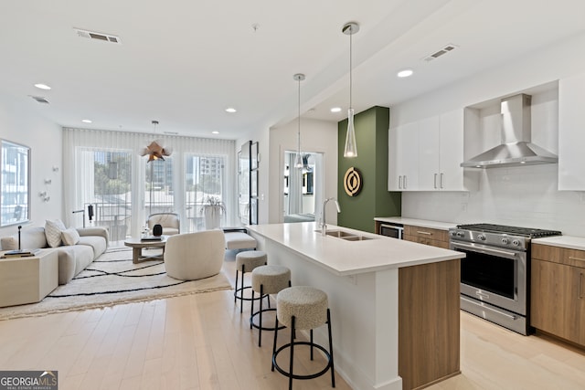 kitchen featuring sink, wall chimney range hood, stainless steel stove, white cabinetry, and an island with sink