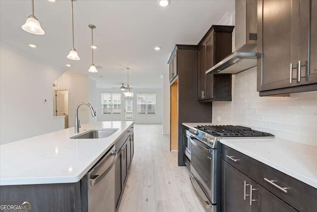 kitchen with sink, hanging light fixtures, stainless steel appliances, wall chimney range hood, and light wood-type flooring