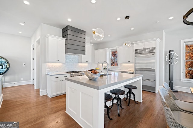 kitchen featuring wood-type flooring, a center island with sink, built in refrigerator, and white cabinetry