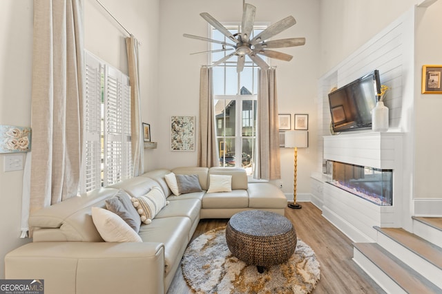 living area with ceiling fan, a high ceiling, baseboards, light wood-type flooring, and a glass covered fireplace