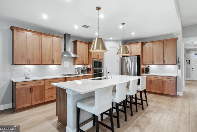 kitchen with sink, wall chimney exhaust hood, an island with sink, light hardwood / wood-style floors, and stainless steel appliances