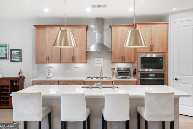 kitchen with stainless steel appliances, light hardwood / wood-style flooring, a kitchen island with sink, and wall chimney exhaust hood