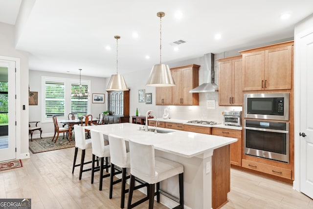 kitchen featuring appliances with stainless steel finishes, a kitchen island with sink, sink, wall chimney range hood, and decorative light fixtures