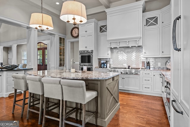 kitchen featuring appliances with stainless steel finishes, beam ceiling, white cabinets, hanging light fixtures, and an island with sink