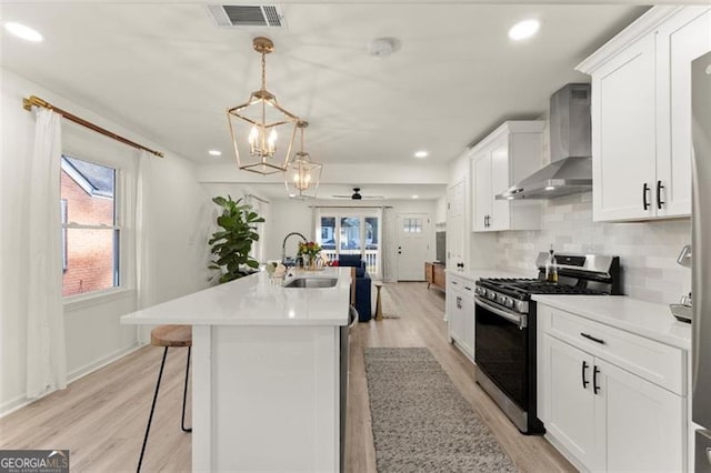 kitchen with sink, wall chimney range hood, stainless steel gas range, pendant lighting, and white cabinets