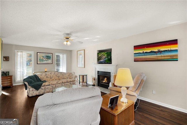 living area featuring a fireplace with flush hearth, baseboards, dark wood-type flooring, and a ceiling fan