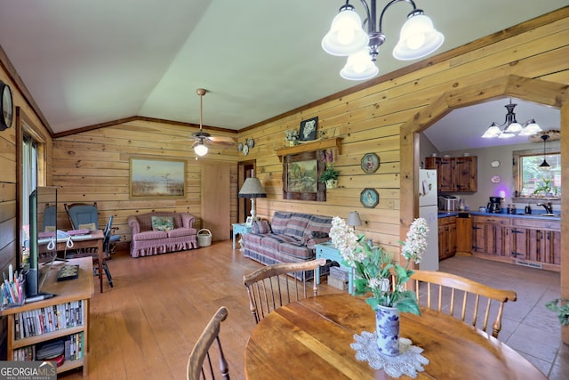 dining room featuring ceiling fan with notable chandelier, lofted ceiling, light hardwood / wood-style flooring, and wooden walls