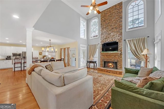 living area with light wood-style flooring, a high ceiling, a stone fireplace, ornate columns, and ceiling fan with notable chandelier