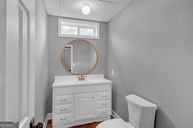 half bathroom featuring toilet, baseboards, a drop ceiling, and vanity