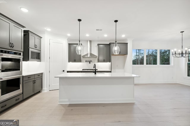 kitchen featuring appliances with stainless steel finishes, wall chimney exhaust hood, light hardwood / wood-style flooring, hanging light fixtures, and an island with sink