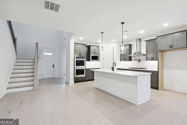 kitchen featuring appliances with stainless steel finishes, a kitchen island with sink, wall chimney range hood, light hardwood / wood-style flooring, and gray cabinets