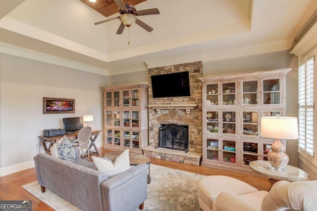 living room with a stone fireplace, light hardwood / wood-style flooring, ceiling fan, ornamental molding, and a tray ceiling