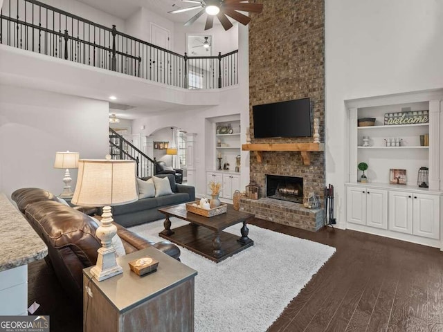 living room featuring built in shelves, ceiling fan, a high ceiling, a brick fireplace, and dark hardwood / wood-style floors