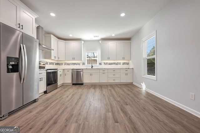 kitchen with white cabinetry, wall chimney exhaust hood, backsplash, appliances with stainless steel finishes, and light wood-type flooring