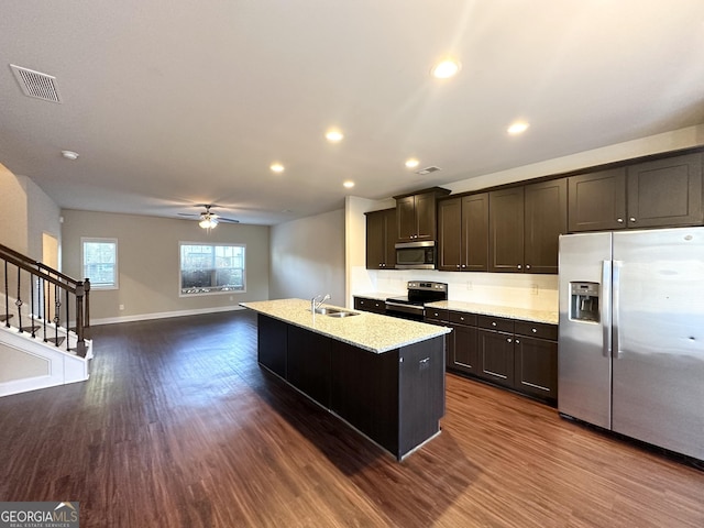 kitchen with dark wood-type flooring, light stone countertops, an island with sink, appliances with stainless steel finishes, and dark brown cabinetry