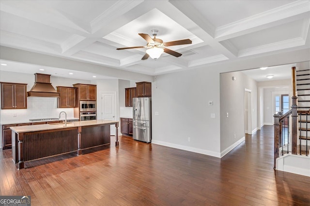 kitchen featuring a breakfast bar area, appliances with stainless steel finishes, a kitchen island with sink, premium range hood, and dark hardwood / wood-style floors