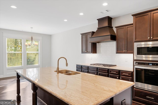 kitchen with sink, custom exhaust hood, an island with sink, stainless steel appliances, and light stone countertops