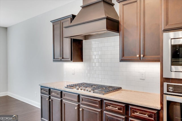 kitchen with decorative backsplash, dark wood-type flooring, light stone counters, stainless steel appliances, and custom range hood