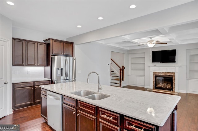 kitchen with sink, stainless steel appliances, coffered ceiling, light stone counters, and a center island with sink