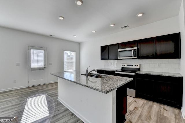 kitchen with a kitchen island with sink, sink, light wood-type flooring, appliances with stainless steel finishes, and light stone counters