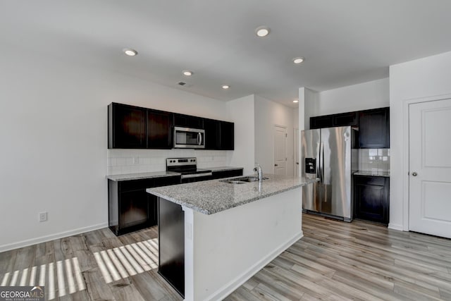 kitchen with decorative backsplash, appliances with stainless steel finishes, light stone countertops, light hardwood / wood-style flooring, and an island with sink