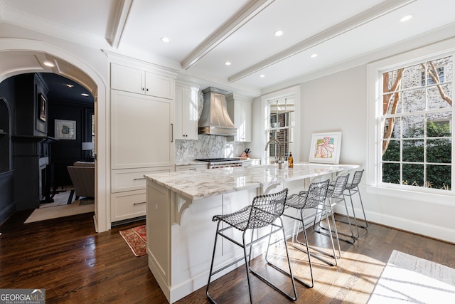 kitchen with white cabinets, a kitchen bar, a kitchen island with sink, and wall chimney exhaust hood