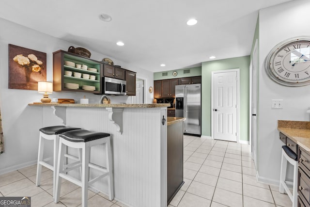 kitchen with a kitchen breakfast bar, light tile patterned floors, stainless steel appliances, light stone countertops, and dark brown cabinets