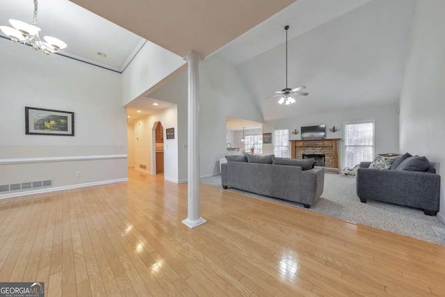 living room with ornate columns, a stone fireplace, ceiling fan with notable chandelier, high vaulted ceiling, and light hardwood / wood-style floors