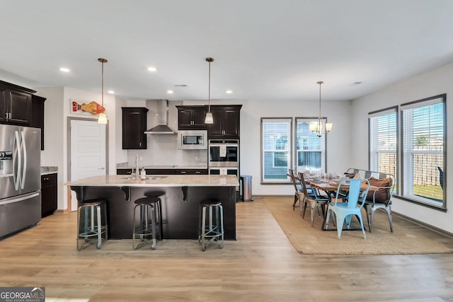 kitchen featuring a breakfast bar, wall chimney range hood, light hardwood / wood-style flooring, an island with sink, and appliances with stainless steel finishes