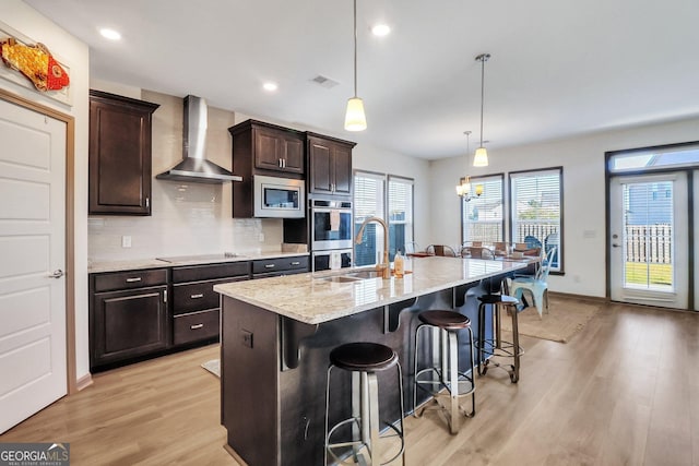 kitchen featuring wall chimney range hood, light hardwood / wood-style floors, decorative light fixtures, a kitchen island with sink, and appliances with stainless steel finishes