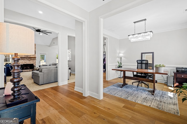 home office featuring ceiling fan with notable chandelier, a stone fireplace, light wood-type flooring, and crown molding