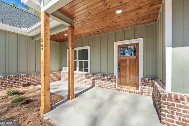 doorway to property with board and batten siding, covered porch, roof with shingles, and brick siding