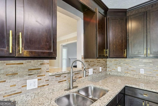 kitchen with backsplash, light stone counters, ornamental molding, dark brown cabinetry, and sink