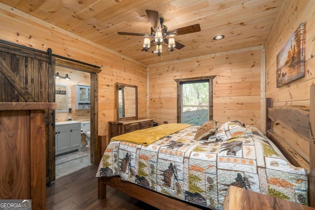 bedroom with dark wood-type flooring, wooden walls, ensuite bath, ceiling fan, and a barn door