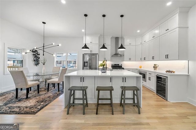 kitchen with hanging light fixtures, wall chimney exhaust hood, appliances with stainless steel finishes, white cabinetry, and beverage cooler