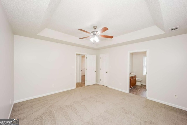 unfurnished bedroom with ceiling fan, a raised ceiling, light carpet, and connected bathroom