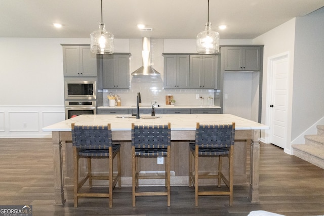 kitchen with gray cabinetry, a sink, appliances with stainless steel finishes, wall chimney exhaust hood, and a center island with sink