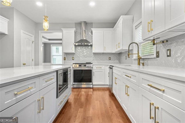 kitchen featuring wall chimney exhaust hood, stainless steel appliances, sink, white cabinetry, and hanging light fixtures