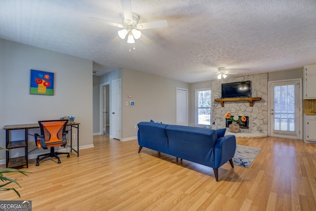 living room with ceiling fan, light hardwood / wood-style floors, a stone fireplace, and a textured ceiling