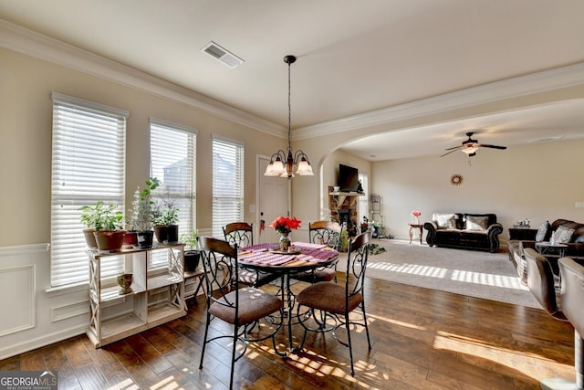 dining area featuring ceiling fan with notable chandelier, a stone fireplace, ornamental molding, and dark wood-type flooring