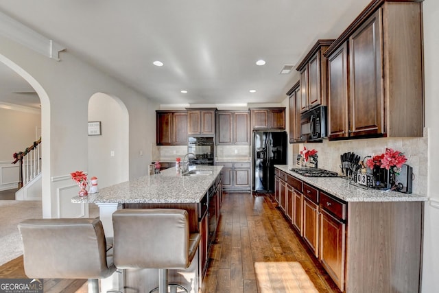 kitchen featuring light stone countertops, backsplash, hardwood / wood-style floors, a kitchen island with sink, and black appliances