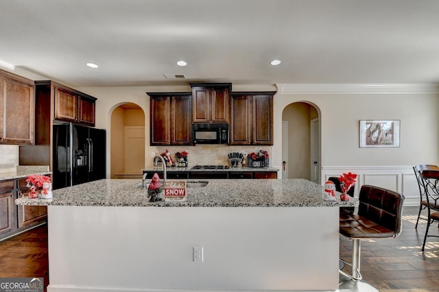 kitchen featuring backsplash, dark wood-type flooring, black appliances, an island with sink, and light stone counters