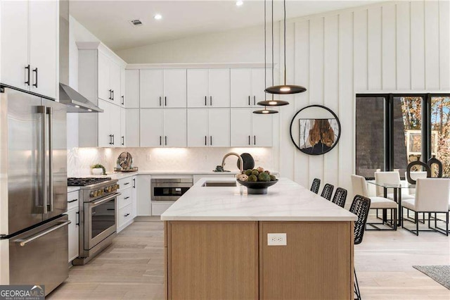 kitchen with high quality appliances, a kitchen island with sink, white cabinets, wall chimney exhaust hood, and light wood-type flooring