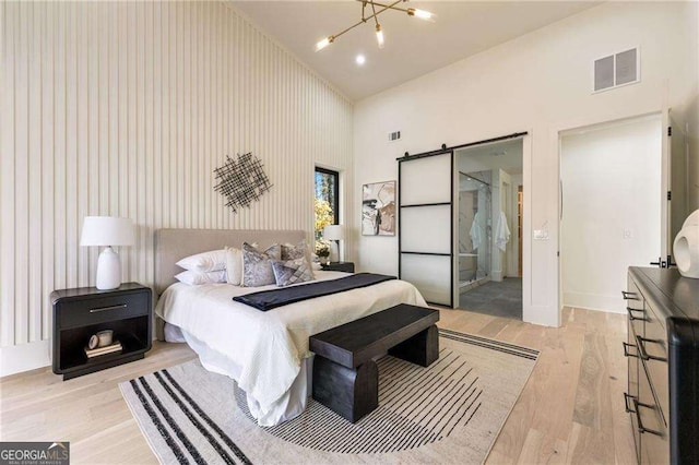 bedroom with high vaulted ceiling, a barn door, light wood-type flooring, connected bathroom, and a notable chandelier