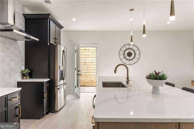 kitchen featuring wall chimney range hood, sink, stainless steel refrigerator with ice dispenser, light wood-type flooring, and decorative light fixtures