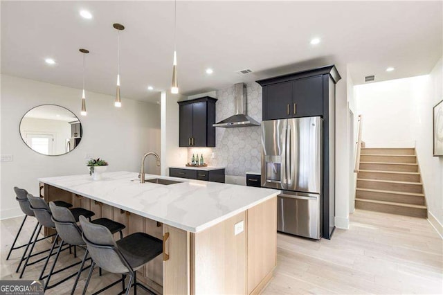 kitchen featuring sink, hanging light fixtures, wall chimney range hood, stainless steel refrigerator with ice dispenser, and a center island with sink