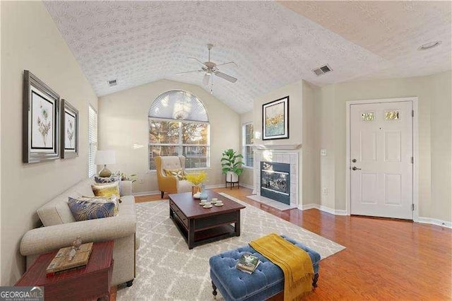 living room featuring light hardwood / wood-style flooring, vaulted ceiling, ceiling fan, a textured ceiling, and a tiled fireplace