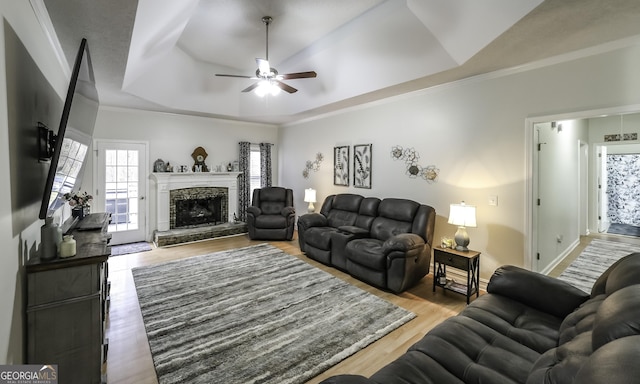 living room with ceiling fan, a stone fireplace, crown molding, wood-type flooring, and a tray ceiling