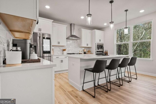 kitchen with appliances with stainless steel finishes, tasteful backsplash, wall chimney exhaust hood, white cabinets, and an island with sink